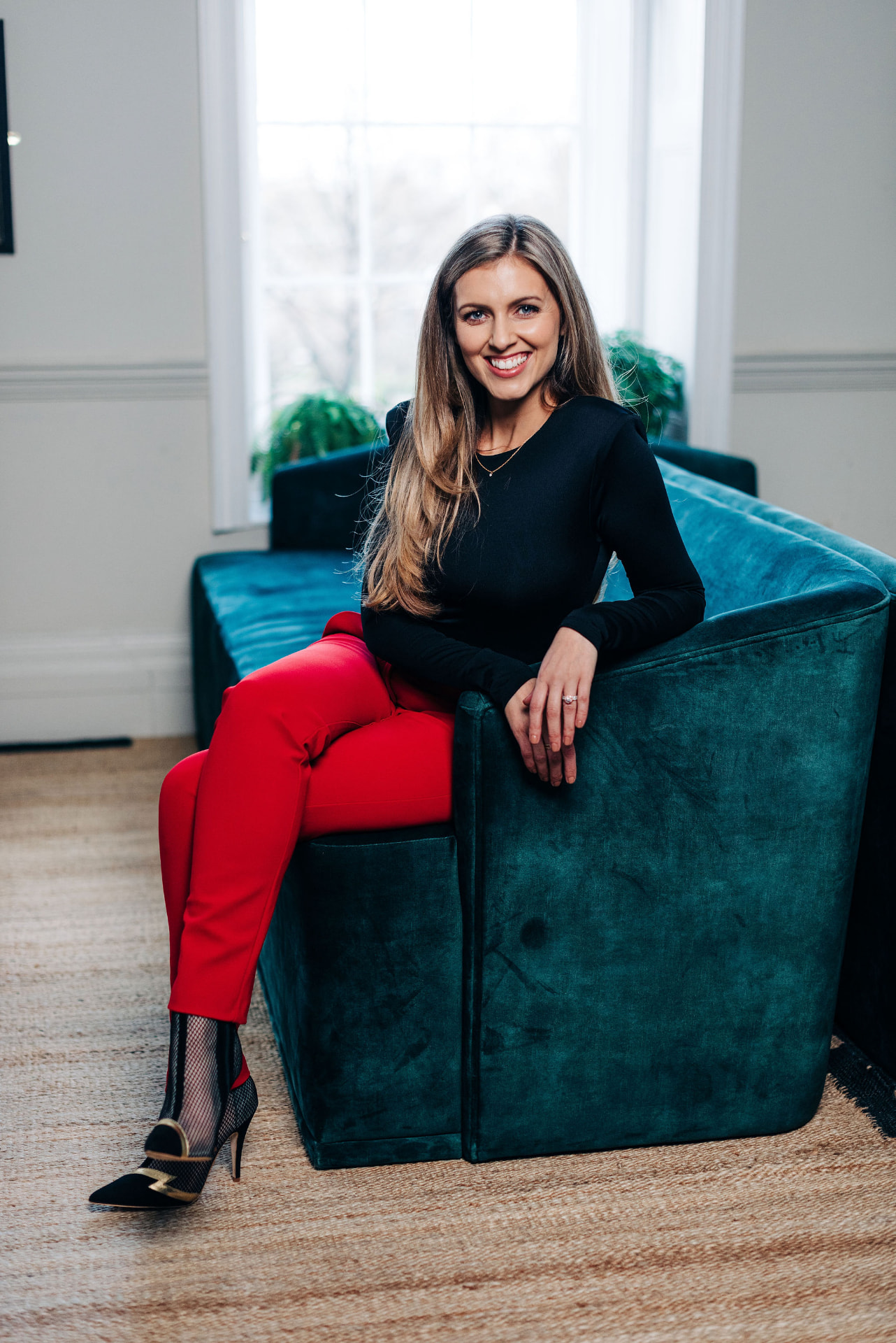 Mikaela Jackson smiling while seated on a teal sofa in red trousers