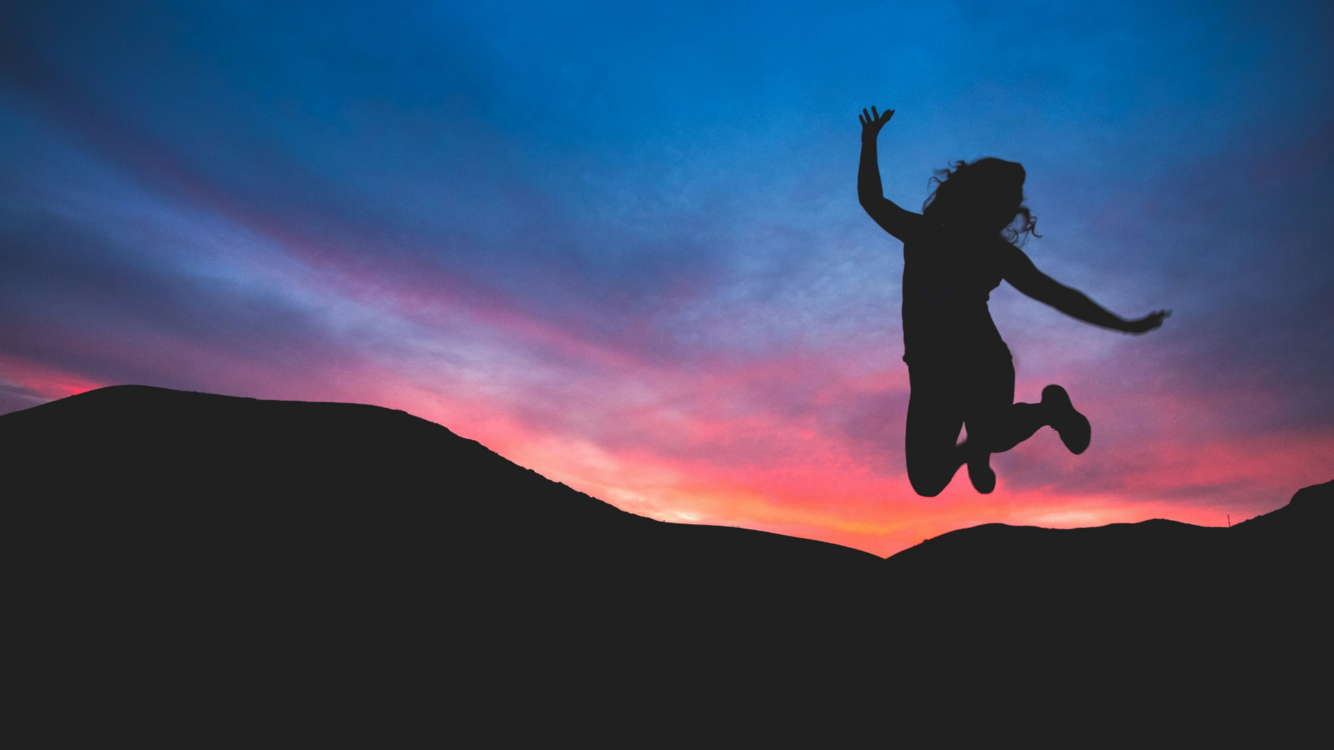 A person leaping in silhouette against a sunset sky over mountain ridges