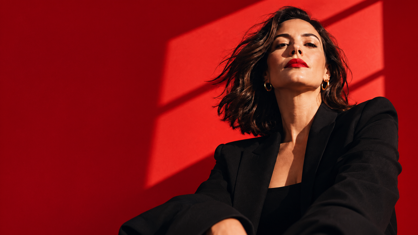 Mikaela Jackson in an oversized black blazer and red lipstick, looking down toward the camera against a red backdrop marked with window shadows